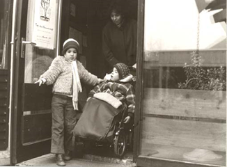 Auf dem Bild hält ein Kind eine Türe auf, wärend eine Frau, ein weiteres
                     Kind im Rollstuhl, aus der Türe nach draußen fährt.