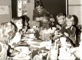 Mehrere Kinder sitzen mit einer männllichern Betreuungsperson an einem
                     Esstisch, an welchem zuvor gegessen wurde.