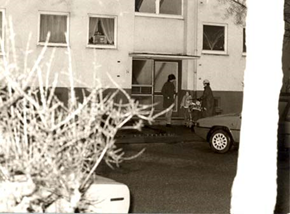 Auf dem Bild ist der Eingang zu einem Gebäude zu sehen, an welchem zwei
                     Personen miteinander sprechen. Vor ihnen steht ein Auto. 
