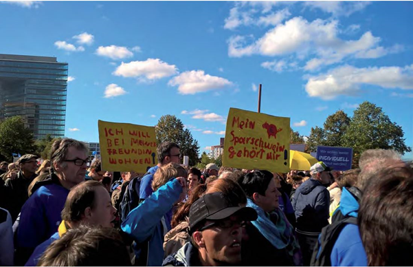 Foto der Protestkundgebung mit mehreren Personen, welche Schilder in die höhe
                  halten. Auf den Schildern steht "Mein Sparschwein Gehört mir!" und "Ich will bei
                  meiner Freundin wohnen!"