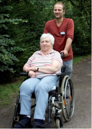 Eine männliche Person schiebt eine ältere Frau im Rollstuhl durch einen Park.
            