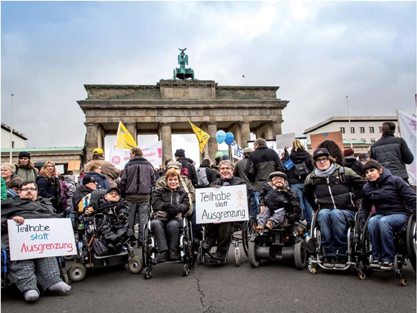 Menschen sitzen in Rollstühlen vor dem Brandenburger Tor und halten Schilder,
                  auf denen "Teilhabe statt Ausgrenzung" steht.