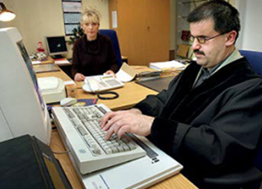 Fotographie eines Mannes der vor dem Computer sitzt. Im
Hintergrund sieht man eine Frau am Telefon.