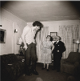 Ein sehr großer Mann steht mit seinen Eltern im
Wohnzimmer. Bild in schwarz-weiß.