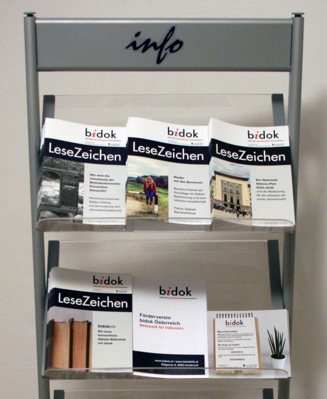 Grauer Broschüren-Ständer mit den zuletzt veröffentlichten bidok LeseZeichen. Am Ständer klebt ein Sticker, darauf steht in dunkelblauer Schrift "Info".