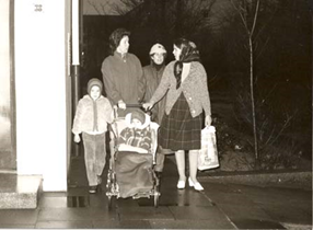 Auf dem Bild sind zwei Kinder und drei Frauen zu sehen, welche in der
                     Nacht vor einem Hauseingang stehen. Ein Kind sitzt in einem Rollstuhl und das
                     Andere steht links daneben. Dahinter die drei Frauen, welche in ein Gespräch
                     vertieft sind.
