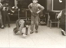 Ein Kind grabbelt auf allen Vieren am Boden und ein weiteres steht
                     daneben.