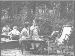 Foto  von Irma Sperling auf dem Spielplatz. Eine Frau und ein Kind
sitzen mit ihr an einem Tisch und sind ins Spiel vertieft. Links sitzen
vier weitere Kinder am Rand eines Sandkastens.